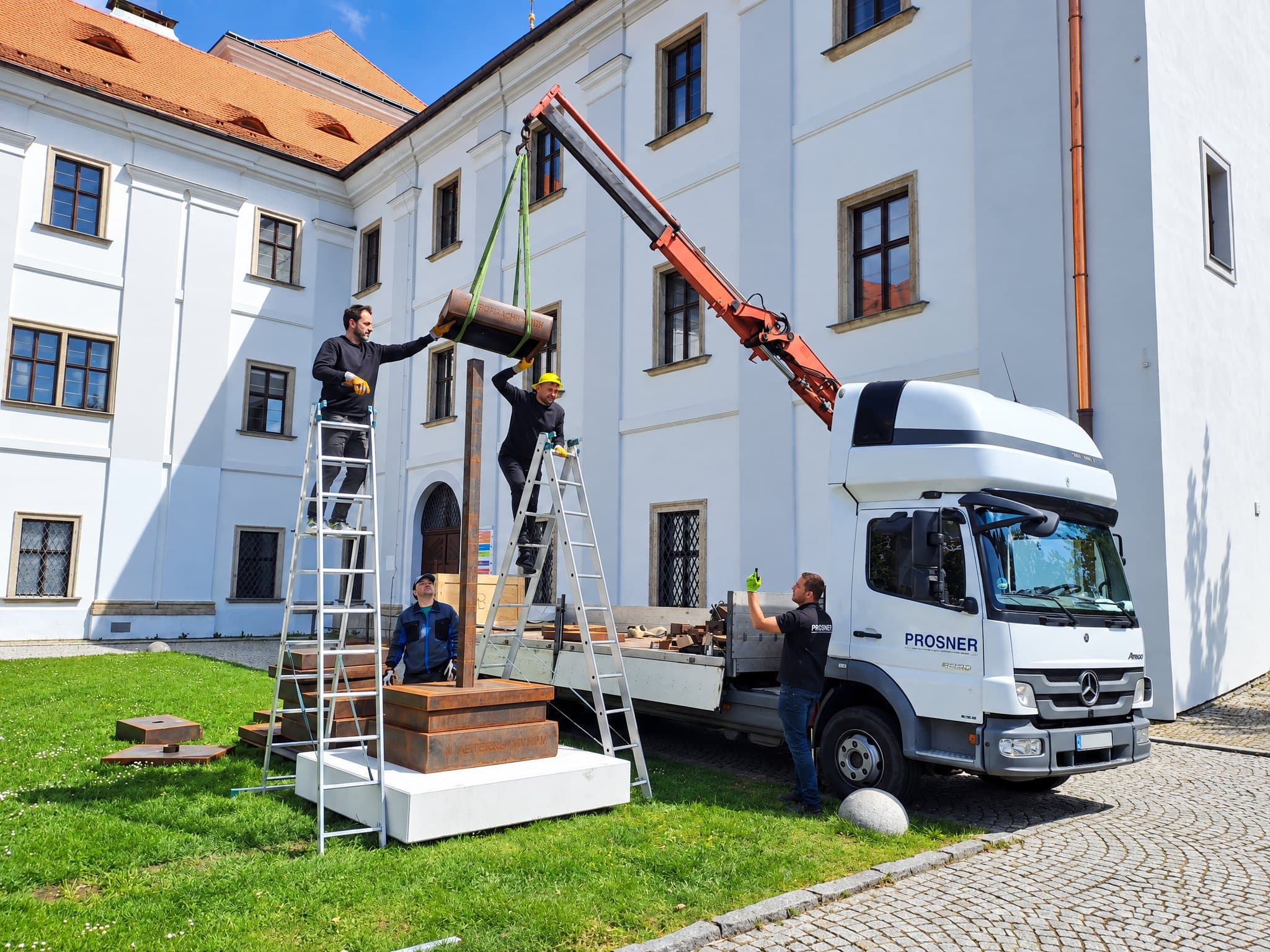 Hydraulická ruka vozu PROSNER drží díl plastiky, který dva muži na žebřících usazují na místo