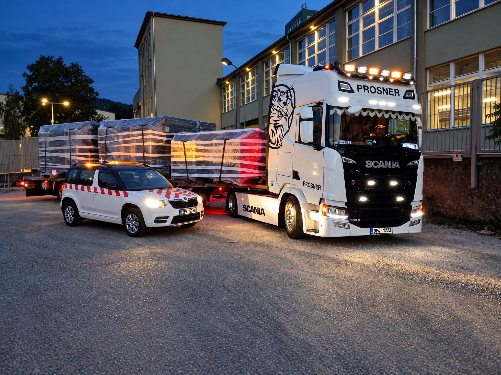 Scania PROSNER s rozsvícenou světelnou rampou veze zaplachtované stroje, vpředu doprovodné vozidlo